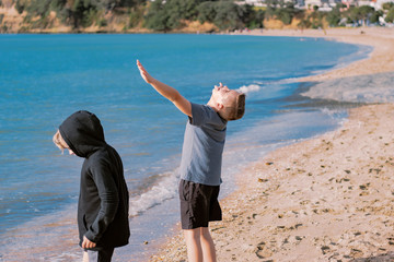 Two young boys, brother, standing on the beach. The older brother has his arms in the air and looking up with his eyes closed; the younger brother has a hooded jacket on.