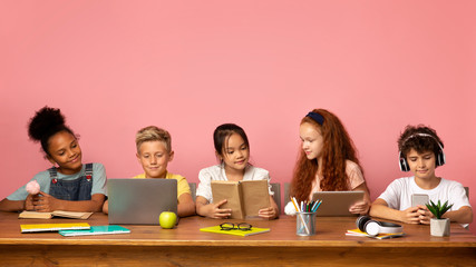 School time. Multiethnic kids with different gadgets and books sitting at table against pink background, empty space