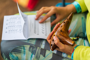 Young Caucasian woman holding a traditional ceramic ocarina in her hand and sheets music papers on her feet, learning how to play on musical notes.