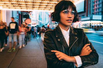 Wall Mural - Contemplative brunette hipster girl in stylish eyeglasses and trendy wear spending evening time in New York standing on street with crossed hands, fashionable female thinking in crowd downtown