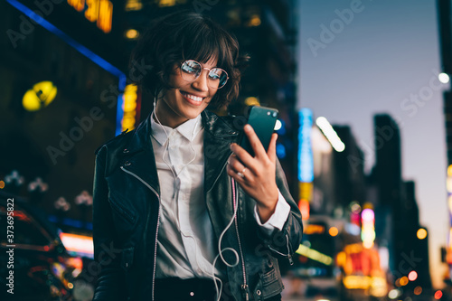 Happy influence blogger in classic glasses and trendy leather jacket reading funny social publication during mobility networking on cellphone, millennial hipster girl listening positive music