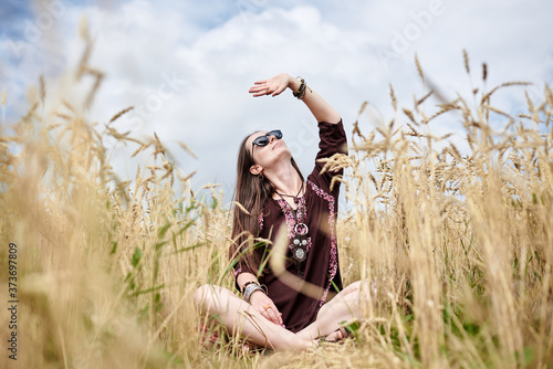 Young brunette hippie woman, wearing boho style clothes, sitting in the middle of wheat field with blue sky, meditating, thinking, relaxing, enjoying. Eco tourism concept. Nature protection.
