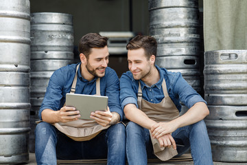 Wall Mural - Two handsome guys in aprons talking and looking at tablet, near metal barrels