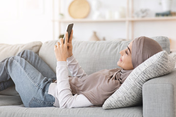 Wall Mural - Happy arabic woman in hijab lying on couch at home using smartphone