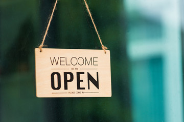 Close up of WELCOME WE ARE OPEN PLEASE COME IN notice sign wood board label hanging through glass door front shop, Business open back to new normal after coronavirus disease concept