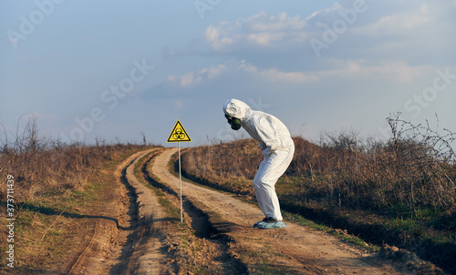 Side view of scientist in suit and gas mask standing by yellow triangle with biohazard symbol on the road under blue sky, suffering from abdominal pain. Concept of ecology, health, biological hazard.