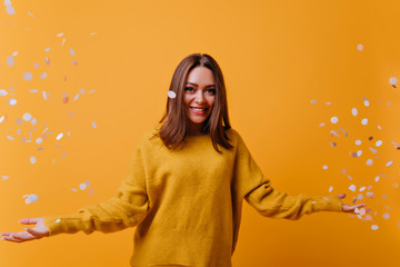 Wall Mural - Happy adorable woman in yellow sweater laughing on bright background. Indoor portrait of appealing girl posing with confetti.