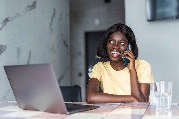 Wall Mural - Smiling african woman using mobile phone while sitting on a kitchen at home