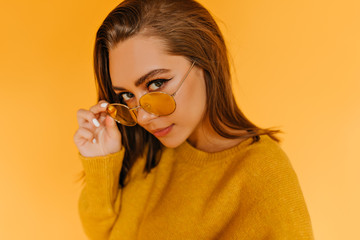 Wall Mural - Fashionable caucasian girl looking through yellow sunglasses. Blissful female model in stylish autumn attire posing in studio.