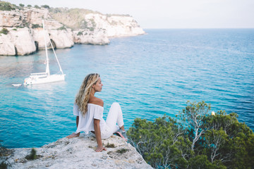 Poster - Carefree tourist spending day for enjoying scenery landscape views of Menorca recreating during adventure journey, Caucasian female solo traveller resting at rocky abyss near berth with yacht