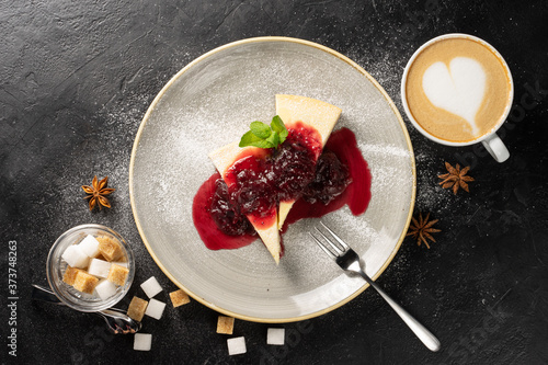 A cup of cappuccino and two slices of cheesecake on a round plate drizzled with strawberry jam and garnished with powdered sugar and mint leaves and scattered with white and brown sugar cubes Poster Mural XXL