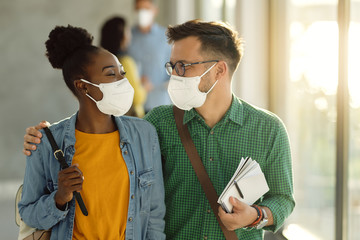 Wall Mural - Happy embraced college couple with protective face masks walking through a hallway.