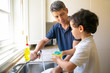 © Mangostar - Lovely little boy helping dad, holding wet sponge and washing plates in kitchen sink. Happy handsome father holding faucet with water and looking at son. Housekeeping and family concept