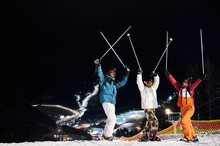 Happy Skiers Team In Winter Jackets And Helmets Raising Ski Poles And Looking At Camera, Standing On Snow-covered Slope With Beautiful Night City On Background. Concept Of Night Skiing And Friendship.