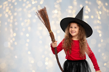 halloween, holiday and childhood concept - smiling girl in costume and witch hat with broom over festive lights background