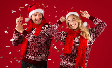 Lovely young couple in sweaters and Santa hats dancing on Christmas or New Year party on red background