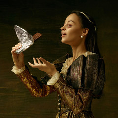 Wall Mural - Medieval young woman in golden vintage clothing as a duchess posing on dark studio background. Concept of comparison of eras, modernity and renaissance. Surreal look with mouth instead eyes.