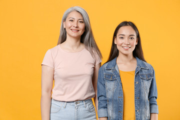 Smiling beautiful attractive family two asian women girls gray-haired mother and brunette daughter in casual clothes posing looking camera isolated on yellow color wall background studio portrait.