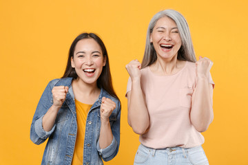 Wall Mural - Happy joyful family two asian women girls gray-haired mother and brunette daughter in casual clothes posing clenching fists doing winner gesture isolated on yellow color background studio portrait.