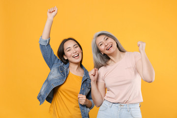 Wall Mural - Cheerful family asian women girls gray-haired mother brunette daughter in casual clothes posing dancing clenching fists rising hands having fun isolated on yellow color background studio portrait.