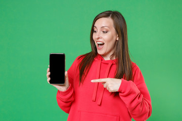 Sticker - Excited young brunette woman in red casual streetwear hoodie pointing index finger on mobile cell phone with blank empty screen mock up copy space isolated on green color background studio portrait.