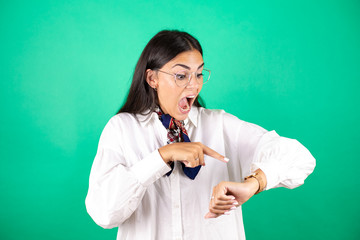 Young beautiful business woman over isolated green background surprised, looking and pointing his watch because it's late