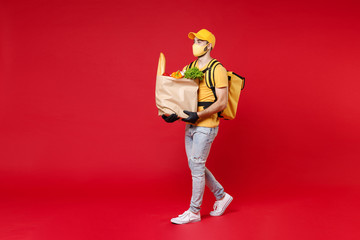 Wall Mural - Full length delivery employee man in yellow cap face mask gloves t-shirt thermal backpack hold paper takeaway food bag products work courier service in quarantine covid-19 isolated on red background.