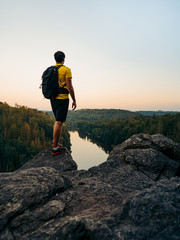 Wall Mural - Young man with backpack and cap on the cliff above river and looking far away. Concept vision, on top of peak mountain and looking forwards, success, competition and leader concept.