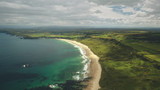 Fototapeta Na ścianę - Aerial view sandy coastline, green grass meadows. Waves crashing out of shore and come back to ocean. Atlantic bay serene scenery in dusk summer daytime cinematic shot