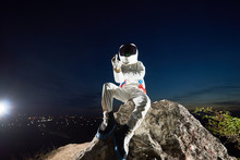 Astronaut Sitting On Top Of Rocky Hill, Doing Rude Hand Gesture While Sitting On Top Of Rocky Hill Under Night Blue Sky. Space Traveler Wearing White Space Suit With Helmet. Concept Of Space Travel