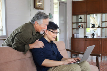 Senior Asian man in retirement age hugging his son from behind while he is working from home using laptop with copy space