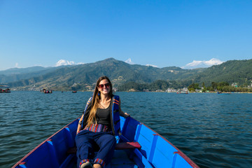 A woman sitting in a blue boat and enjoying a tour across Phewa Lake in Pokhara, Nepal. Behind her there are high, snow capped Himalayas with Mt Fishtail (Machhapuchhare) between them. Relaxation