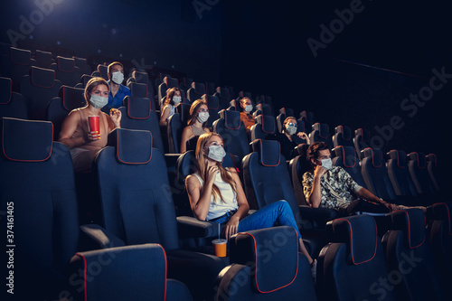 Cinema, movie theatre during quarantine. Coronavirus pandemic safety rules, social distance during movie watching. Men and women wearing protective face mask sitting in a rows of auditorium, eating