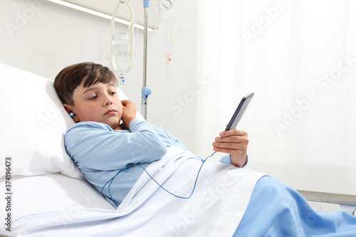 Fotografía child in hospital bed using smartphone surfs the internet wearing earphones