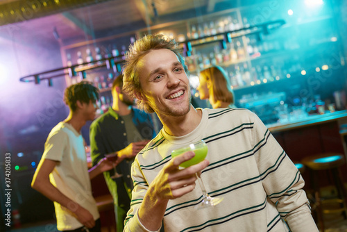 Entertainer. Caucasian young man looking aside while posing with a cocktail in his hand and friends chatting, having drinks at the bar counter in the background