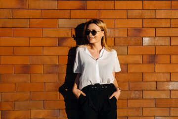 Young stylish woman wearing sunglasses, white shirt and black pants, standing on a brick wall background outdoors, smiling.