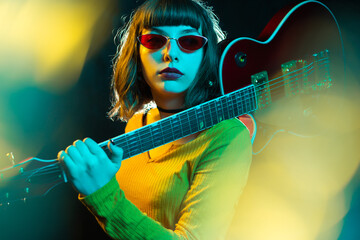 Stylish young hipster woman with curly hair holding red guitar on shoulder in neon lights. 90s style concept.