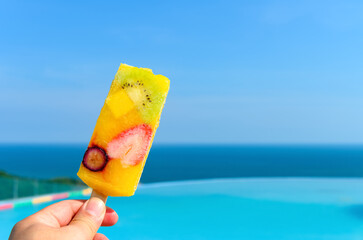 Wall Mural - hand holding a fresh fruit popsicle in front of a swimming pool with the ocean at the far end