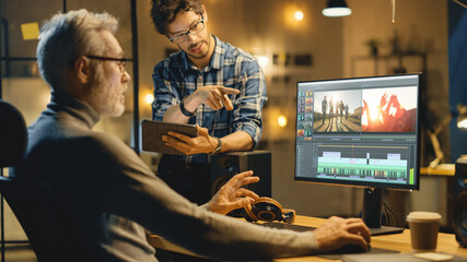 Evening in Creative Office: Professional Middle Aged Videographer Works on Desktop Computer, Has Conversation with Younger Specialist who Uses Tablet Computer. Modern Video Developers Office