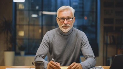 Canvas Print - Portrait of the Handsome and Successful Middle Aged Bearded Businessman Working at His Desk Using Laptop Computer. Smiles at Camera. Working at Night from Cozy Home Office / Studio with Window