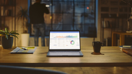 Shot of a Laptop on a Desk with Statistics Graph, Chart and Various Data, Showing Company Growth and Success. In the Background Businessman Drinks from a Cup Looks at the Night City out of the Window