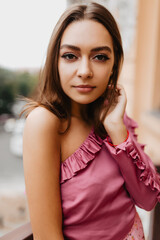 Wall Mural - Pensive girl calmly looks into camera, posing for street photo. Cute model in stylish top with frills straightens her hairdo