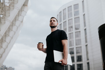 young handsome man walking down the street, carrying his laptop while drinking coffee on the go, lunch break, busy days, multitasking