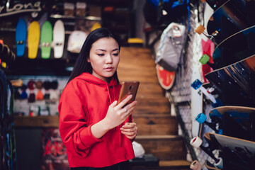 Millennial female teenager using cellphone for browsing web store and compare prices during shopping day for choosing youthful skateboard, Chinese generation Z making banking payment for purchase