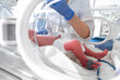 Photo of a premature baby in incubator. Focus is on his feet. Nurse in blue gloves is using the feeding tube for feeding premature baby. Neonatal intensive care unit