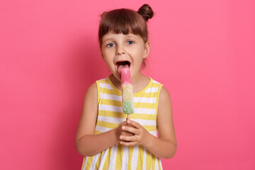 Adorable preschool female child holding sorbet and looking at camera. Cute little girl licking fruit ice-cream, widely opens mouth, standing against pink wall,