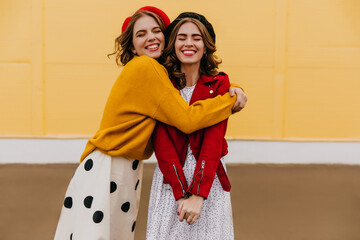 Stylish french girl embracing best friend on yellow background. Outdoor portrait of brunette ladies smiling to camera.