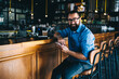 © BullRun - Cheerful handsome mature man in casual wear and spectacles sitting near bar indoors with smartphone, happy prosperous 40 years male owner of small business satisfied with income from cafetaria