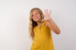 © Roquillo - Little blond girl wearing yellow dress over white background Waiving saying hello happy and smiling, friendly welcome gesture.