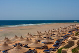Fototapeta Do akwarium - View of the sandy beach on which there are natural umbrellas, sunbeds. In the background is the sea and pontoons to enter the sea.